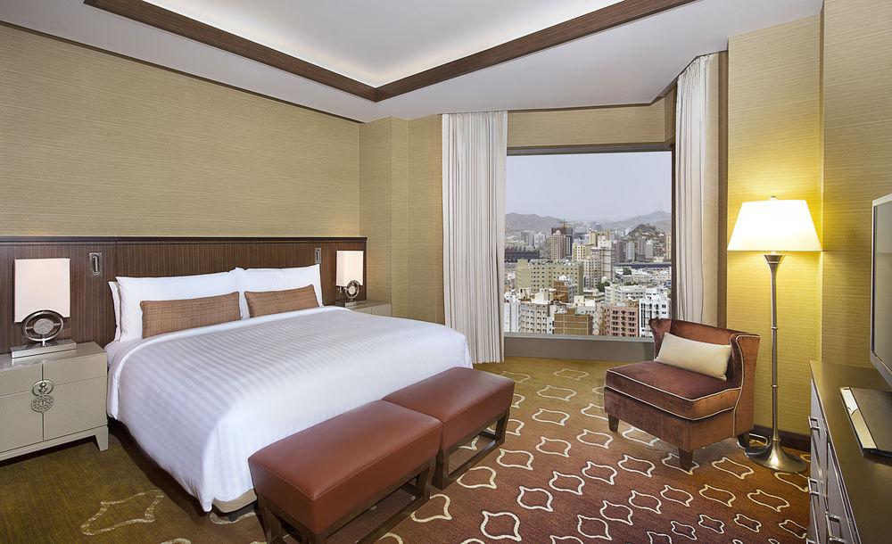 자발 오마르 메리어트 호텔, 마카 메카 외부 사진 The photo shows a modern hotel room featuring a large bed with white linens and decorative cushions. There are two brown leather footstools at the foot of the bed. Next to the bed is a stylish armchair in a complementary color. The room has large win